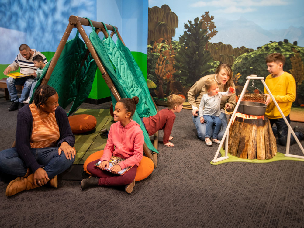 Two families sitting and reading near a green tent. One family gathered around a pretend campfire.